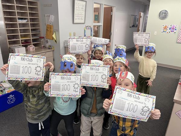 students smiling and holding up 100 Club worksheets while wearing glasses and crowns shaped like the number 100